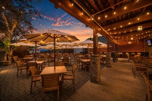 Eine Terrasse im Freien mit Tischen und Stühlen, beschattet durch Sonnenschirme und beleuchtet mit Lichterketten.