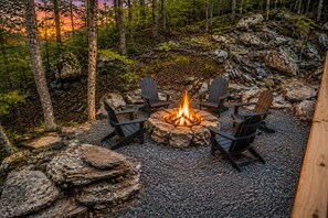 Ein Lagerfeuer, umringt von Adirondack-Stühlen, inmitten eines Waldes.