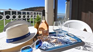 A table set with a bottle of wine, glasses, a hat, and sunglasses on a patio with a view of a hill and a white fence. - L'Esquinade (F4 218) by Interhome (Agay Village Cap Esterel)