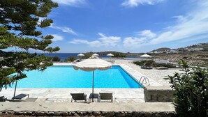Una piscina amb gandules i ombrel·les, envoltada de murs de pedra i vegetació, amb vista al mar i edificis allunyats.