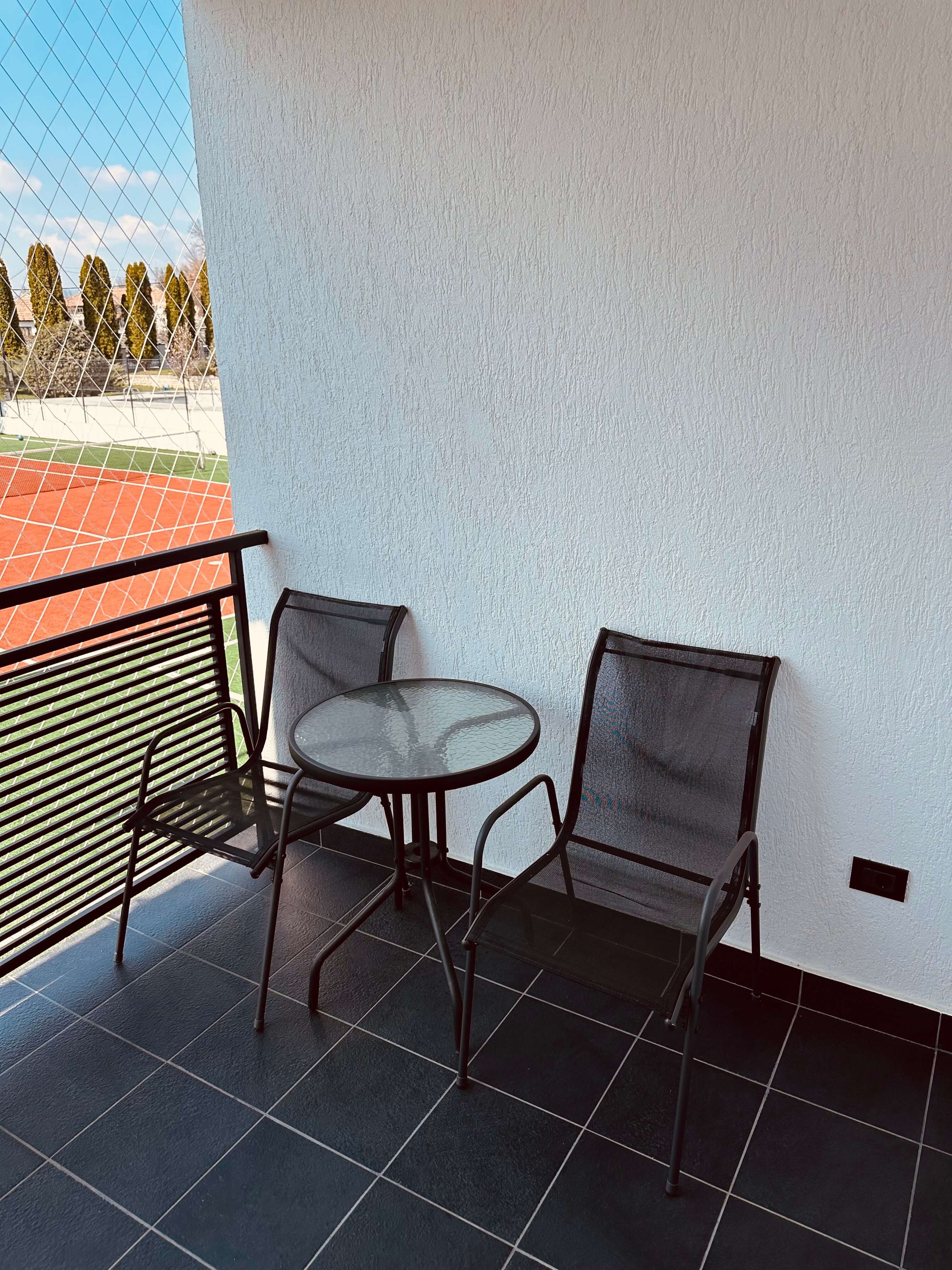 A balcony with a round table and two chairs, a metal railing, and a view of a sports field.
