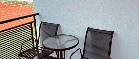 A balcony with a round table and two chairs, a metal railing, and a view of a sports field.