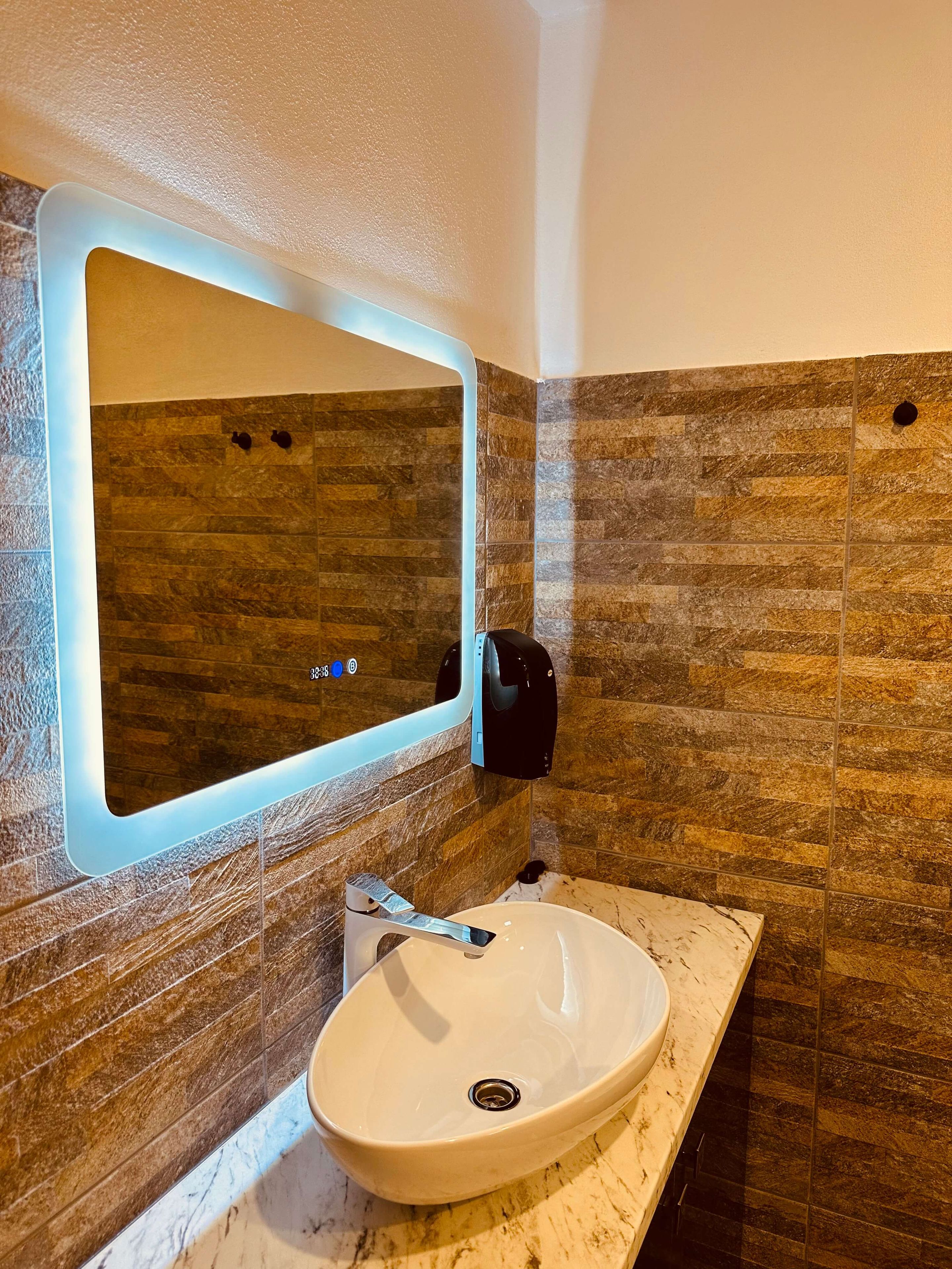 A bathroom with a marble sink, a wall-mounted mirror with a light, and a soap dispenser.