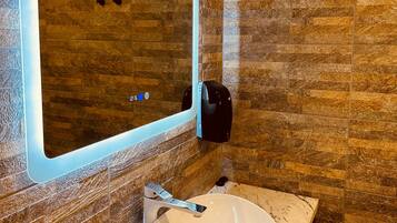A bathroom with a marble sink, a wall-mounted mirror with a light, and a soap dispenser.