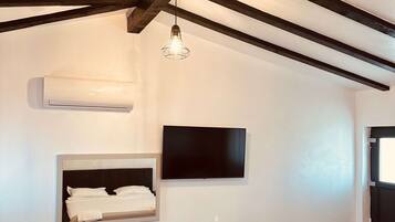 A modern hotel room with exposed wooden beams, a flat-screen TV, a built-in desk with a mirror, and a ceiling light fixture.