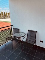 Un balcón con una mesa redonda y dos sillas, barandilla metálica y vista a un campo deportivo.