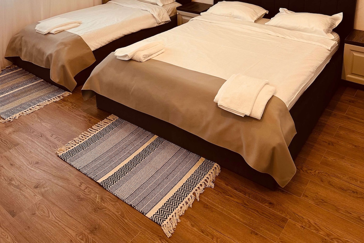 Habitación de hotel con dos camas, suelo de madera, alfombra estampada y techo con vigas a la vista.
