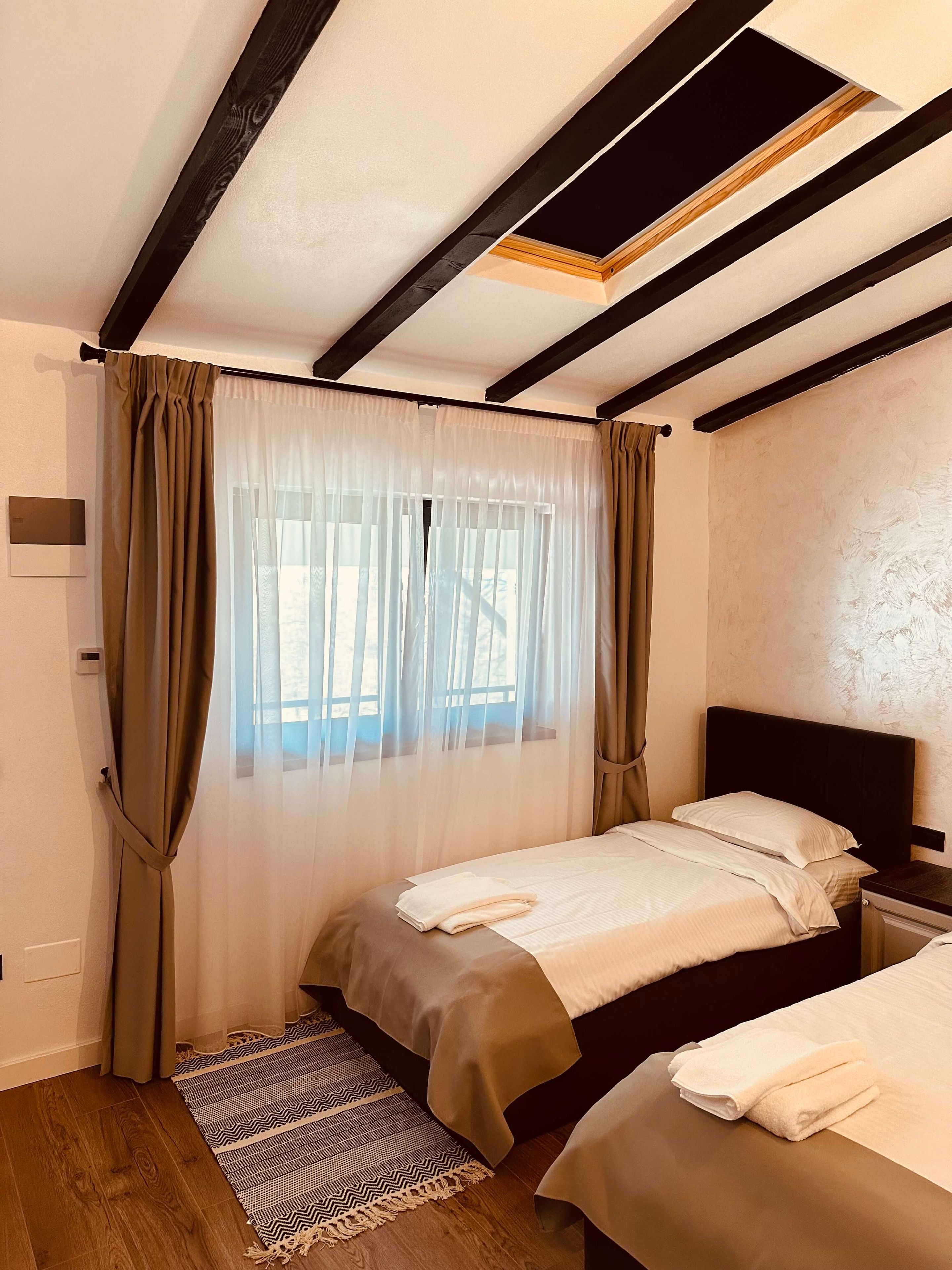 A bedroom with a bed, a bedside table, a window with sheer curtains, and a patterned rug.
