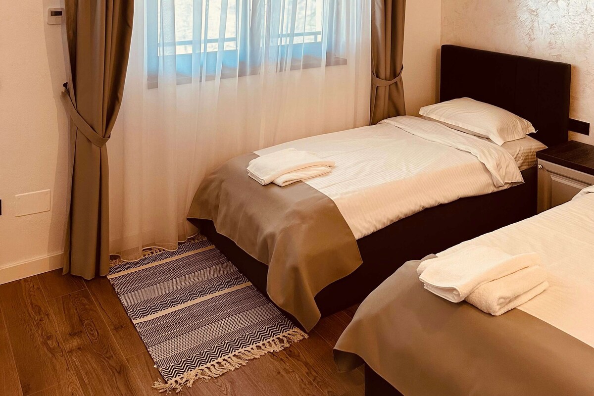 Un dormitorio con cama, mesita de noche, ventana con cortinas translúcidas y una alfombra estampada.
