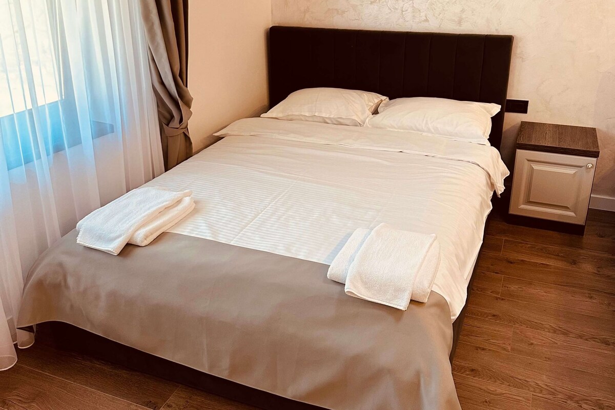 Una cama bien hecha con sábanas y toallas blancas, una mesita de noche de madera con cajón, y una ventana con cortinas traslúcidas.
