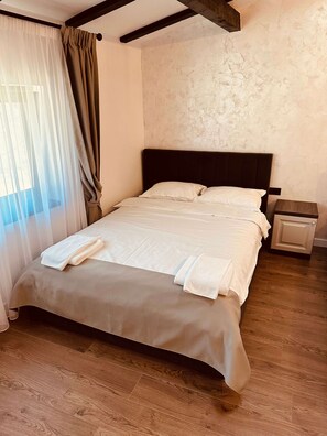 Una cama bien hecha con sábanas y toallas blancas, una mesita de noche de madera con cajón, y una ventana con cortinas traslúcidas.