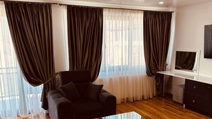 A modern living room with a black sofa, a glass coffee table, wooden flooring, and large windows with dark curtains. - Casa Yda (Horezu)