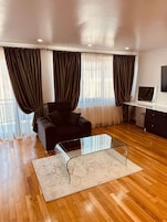 Una sala de estar moderna con un sofá negro, una mesa de centro de vidrio, piso de madera y ventanas amplias con cortinas oscuras.
