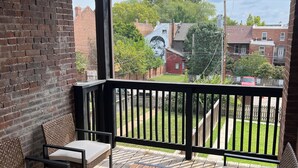 Eine Holzterrasse mit zwei gepolsterten Stühlen und einem Tisch, einem hängenden Laternen und Blick auf eine Wohngegend mit Häusern und einem Basketballplatz.