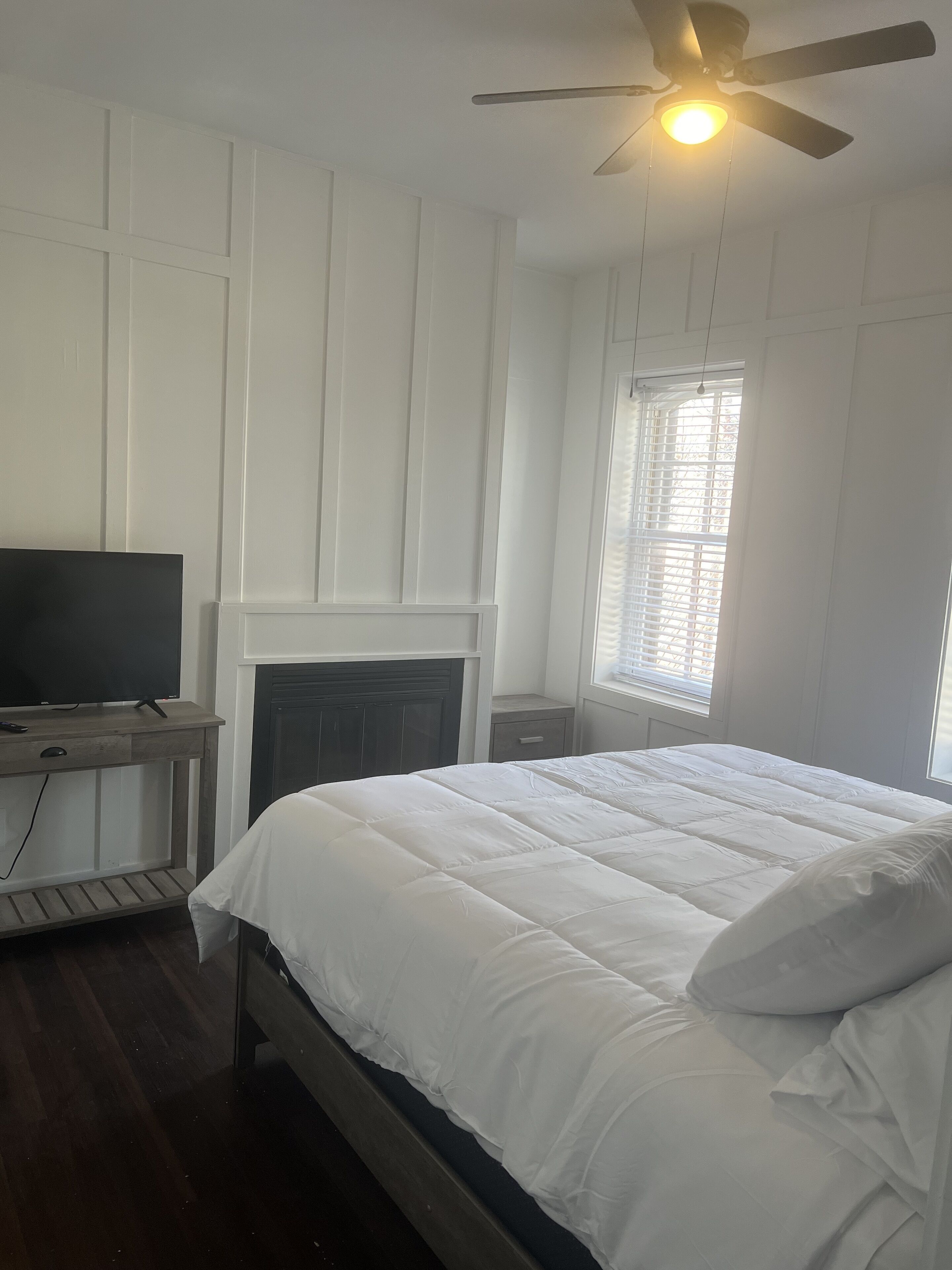 A bedroom with a bed, a television on a wooden table, a ceiling fan, and a window with blinds.