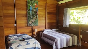 A wooden cabin room with two beds, a nightstand, a lamp, and a painting of parrots and a sloth. - Quirquincho House (Tambopata)