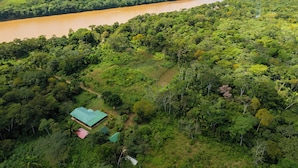 An aerial view of a lush green landscape with a river, dense forest, and a small building with a green roof. - Quirquincho House (Tambopata)