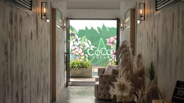 Un couloir avec un canapé et une plante, qui mène à une pièce où est affiché un panneau indiquant « Coco House ».
