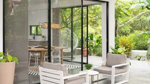Un espace extérieur moderne aménagé avec des sièges, doté d’une porte vitrée donnant sur une cuisine et un coin repas, le tout entouré d’une végétation luxuriante.