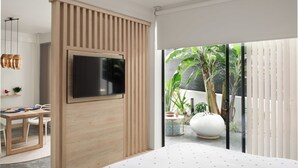 Un salon moderne avec un téléviseur à écran plat fixé sur un panneau en bois, un coin repas avec une table et des chaises, et une grande fenêtre donnant sur un jardin.