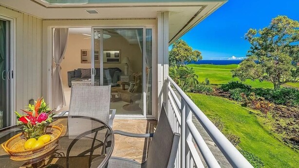 A balcony with a glass door leading to a living area, a table with a basket of fruit, and a view of a green landscape.