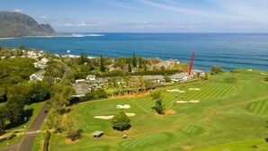 An aerial view of a golf course with a clubhouse, a road, and a view of the ocean and mountains in the background.