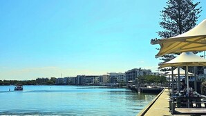 Een promenade aan het water met een betonnen wandelpad, een overdekt zitgedeelte en een uitzicht op het water en gebouwen op de achtergrond.