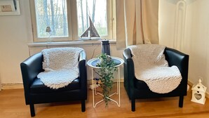 Una habitación con dos sillones de cuero negro, cada uno con una manta blanca. Hay una pequeña mesa redonda con una planta en el centro. Se ve una ventana grande con cortinas, y una lámpara negra cuelga de la pared.