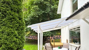 Un patio con mesa y sillas bajo una pérgola, rodeado de follaje y una casa.