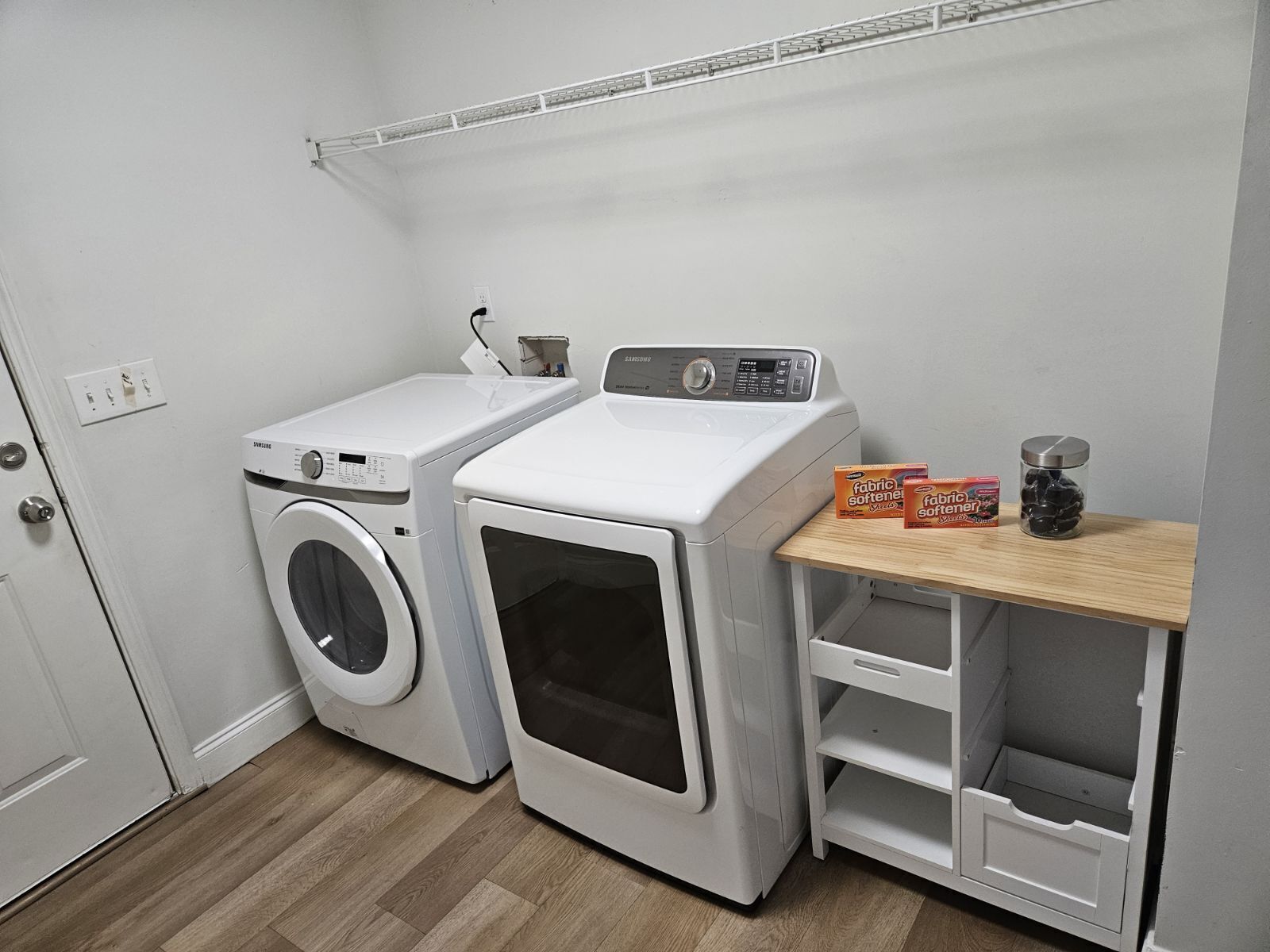 A laundry room with two white washing machines, a shelf with fabric softener boxes, and a jar of black pebbles.