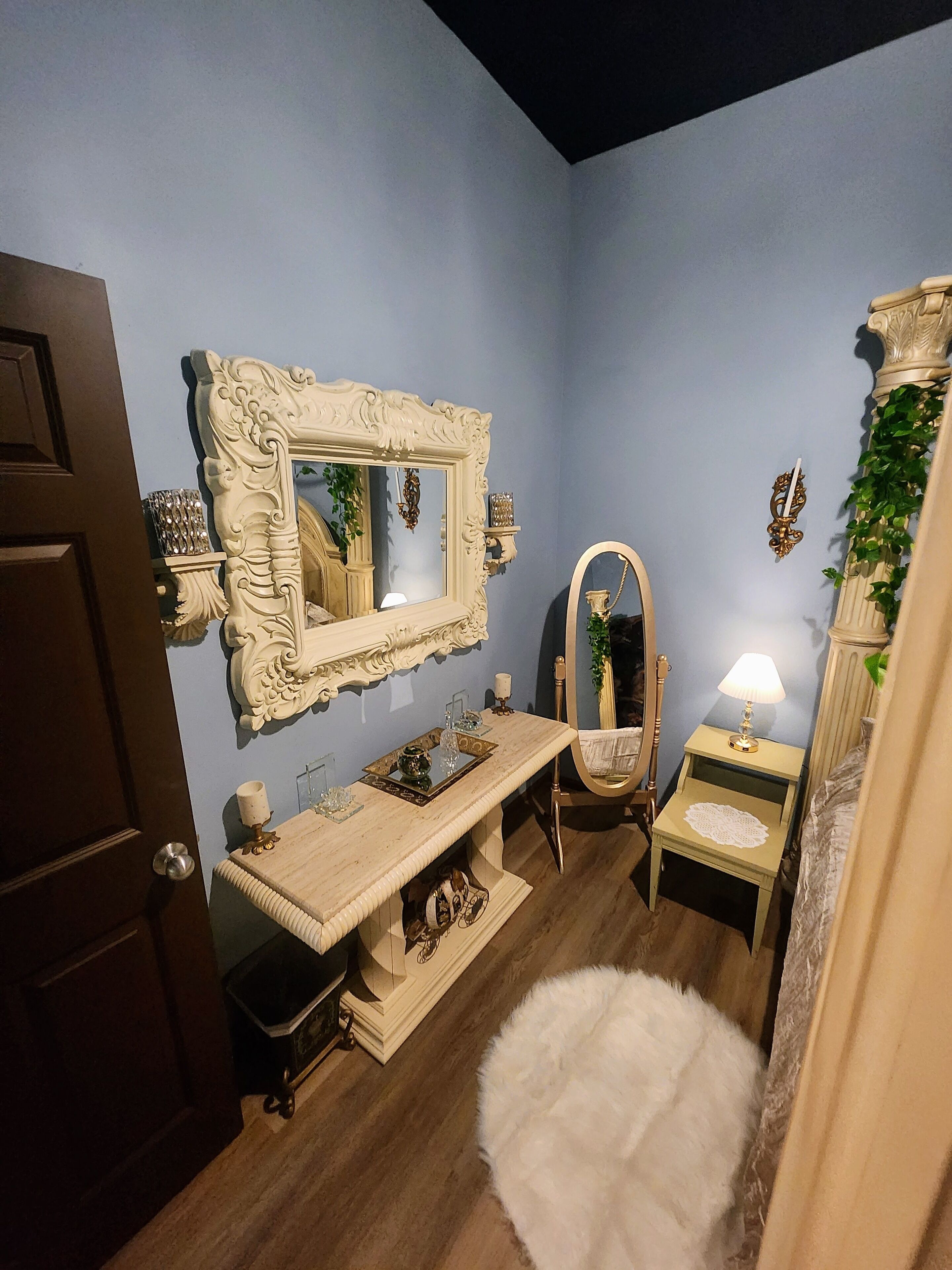 A bathroom with a vanity, a large mirror, a small table with a lamp, and a door.