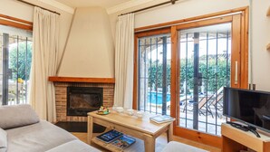 Un salon confortable avec une cheminée, un téléviseur et une vue sur une piscine et la verdure environnante.