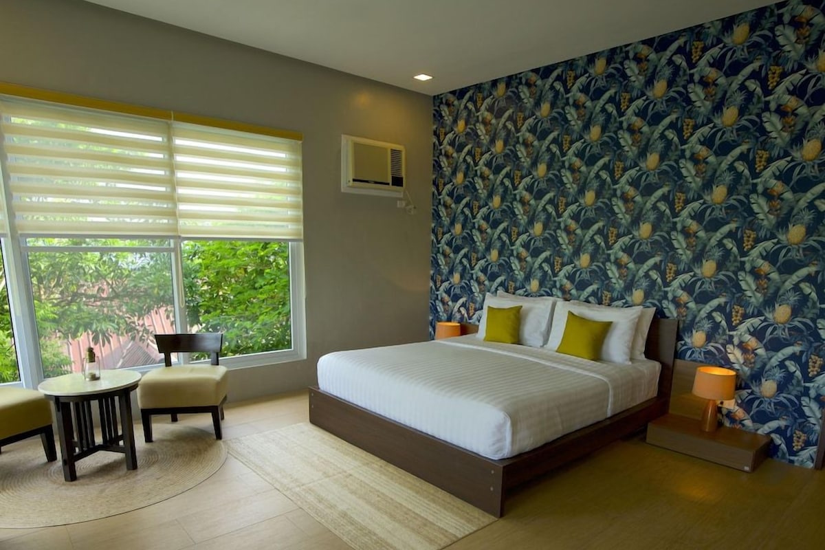A bedroom with a large bed, a small round table, two chairs, a window with blinds, and a patterned wall.