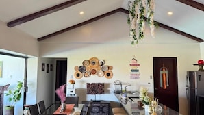A modern kitchen with a central island, dark wooden beams, hanging plants, and decorative wall art.