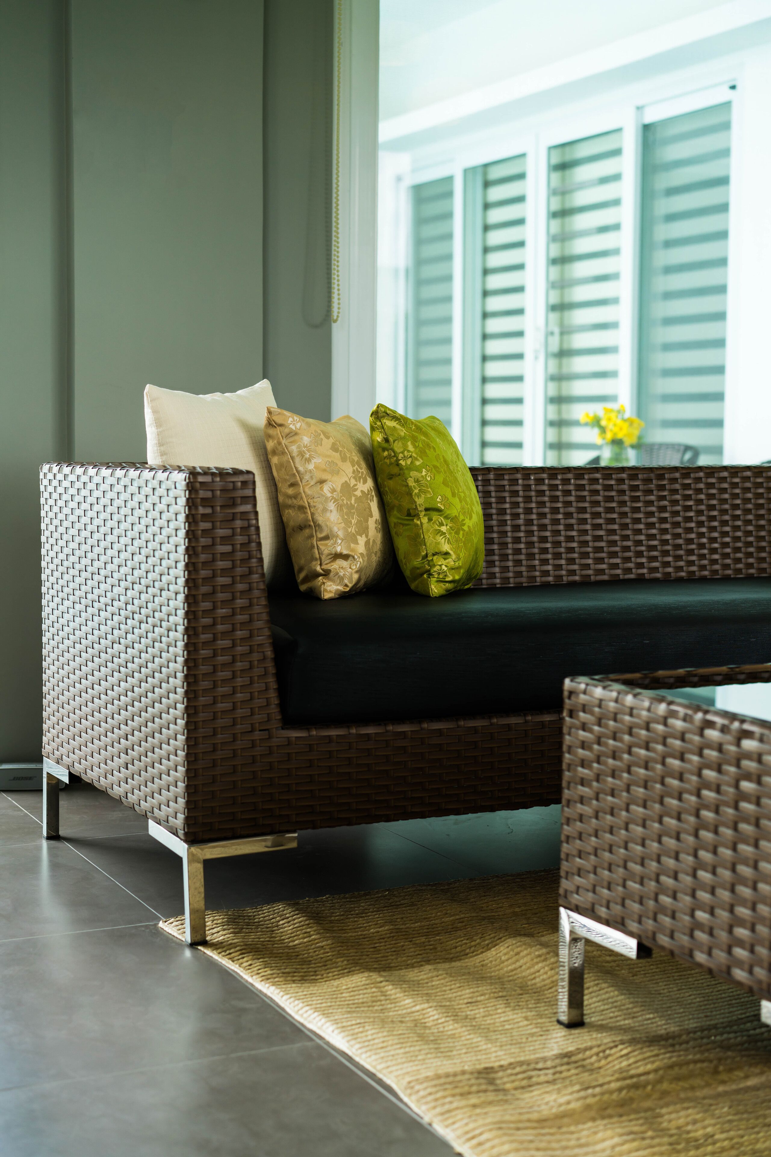 A wicker sofa with cushions, a woven rug, and a view of a window with shutters.