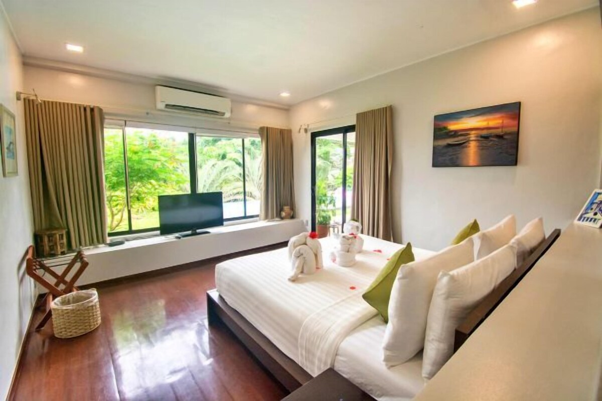 A hotel room with a bed, a TV, a window with curtains, a painting, and a wooden floor.