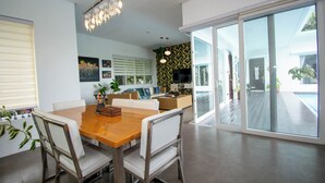 A modern dining area with a wooden table and white chairs, a sofa, and a TV in the background.