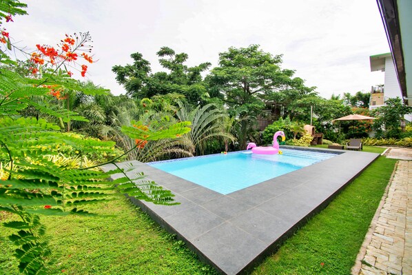 A rectangular swimming pool with a dark tiled edge, surrounded by lush greenery and trees.