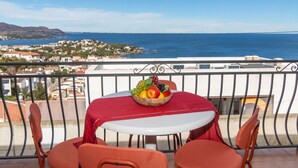 Ein Balkon mit Tisch für zwei Personen, ausgestattet mit einer Obstschale und einer roten Tischdecke. Der Blick umfasst eine Küstenlandschaft mit Gebäuden und einer Brücke.