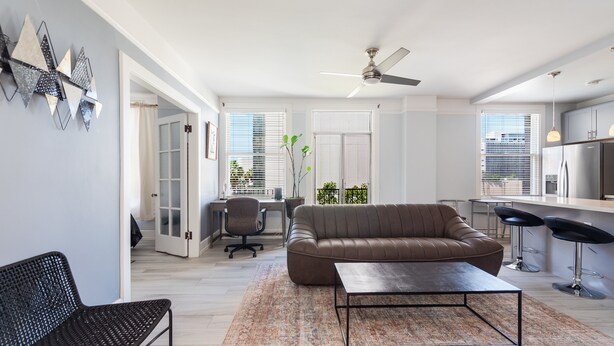 A modern living room with a brown leather sofa, a black metal coffee table, and a patterned rug. There is a black office chair, a desk with a computer, and a ceiling fan. - Executive-next to Convention Center + Free Parking (Long Beach)