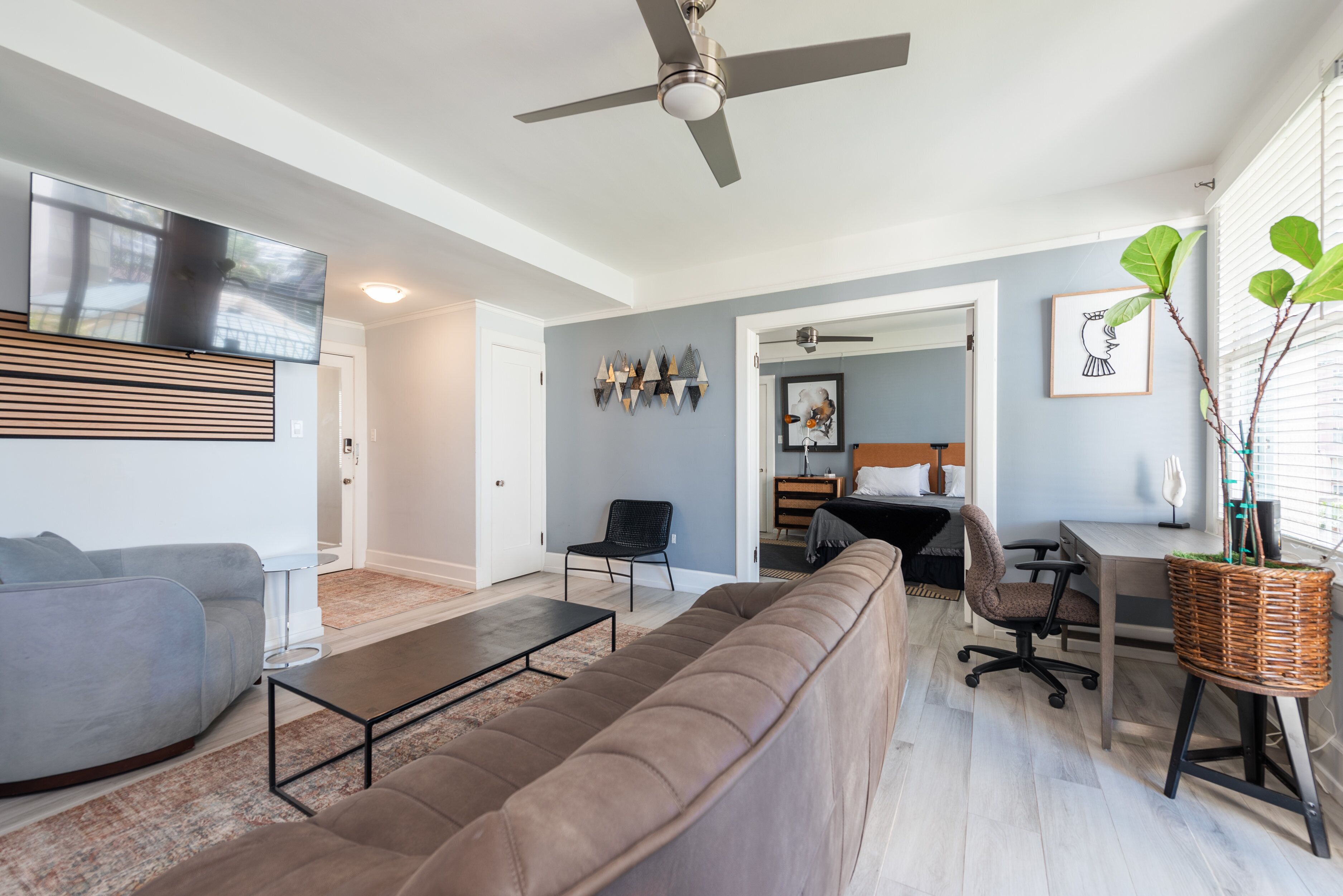 A modern living room with a sofa, coffee table, and a TV mounted on the wall. There is a ceiling fan, a dining area with a table and chairs, and a small office space.