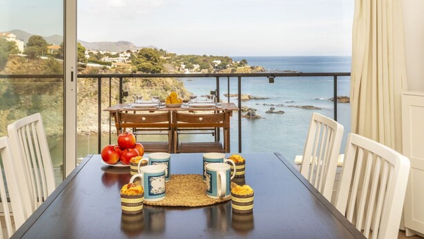 Mesa de jantar posta para dois com vista panorâmica do oceano, um sousboche de vime e um prato de frutas.