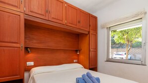 A bedroom with a bed, wooden wardrobe, and a window with a view of a tree and a car. - Mar i Muntanya II by Interhome (Llançà)