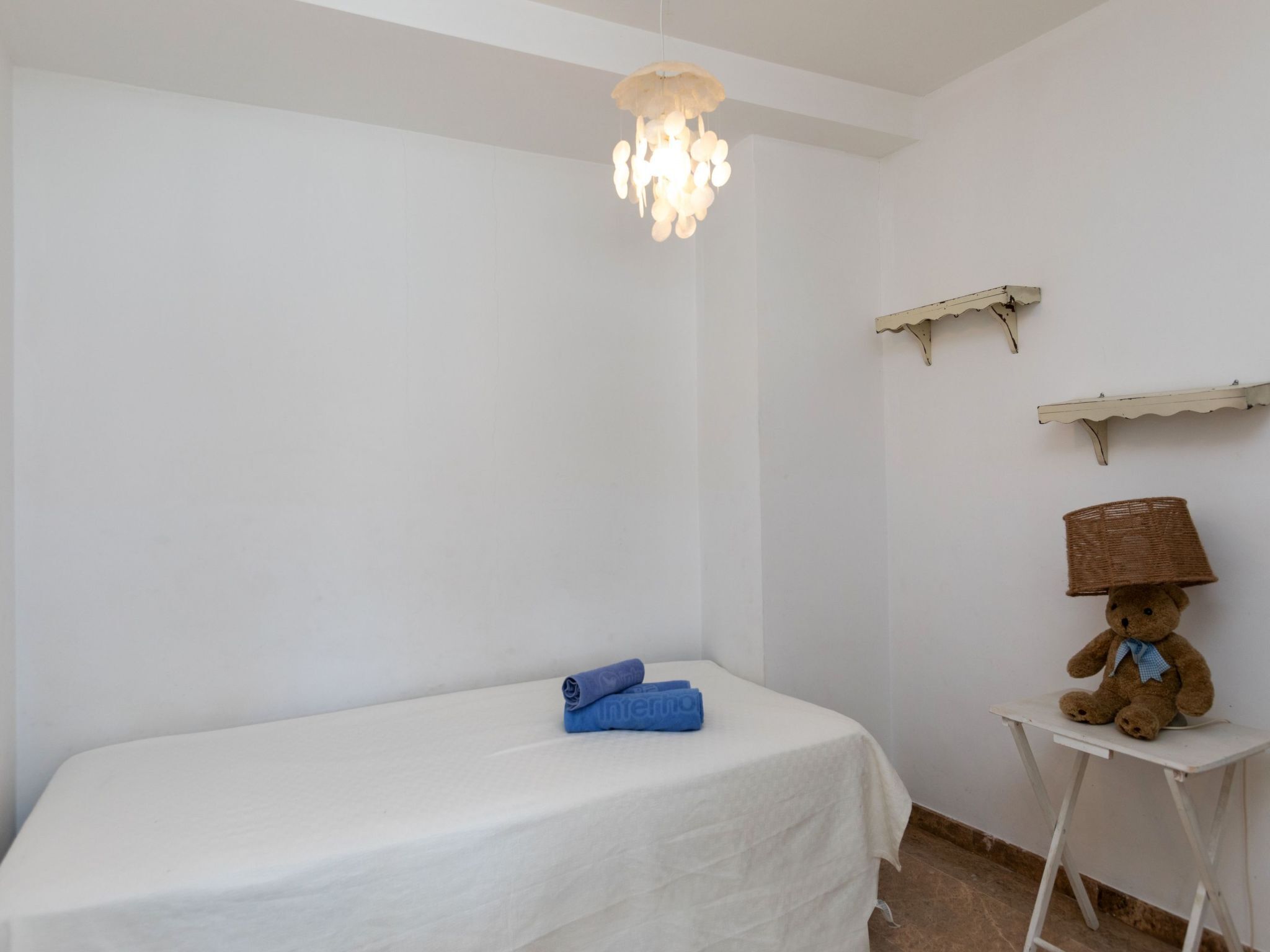 A small, white-walled room with a single bed, a small table, and a teddy bear on the table.