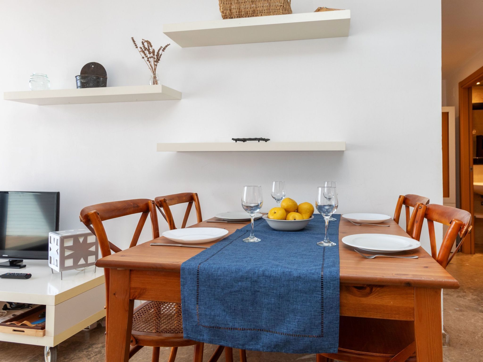 Un'area pranzo con un tavolo di legno, tovaglia blu e sedie. Sul tavolo ci sono piatti bianchi, bicchieri e una ciotola di limoni.