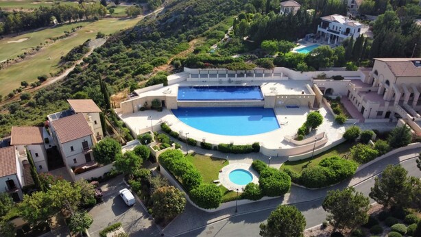 An aerial view of a resort with a large central pool, multiple buildings, and surrounding greenery.
