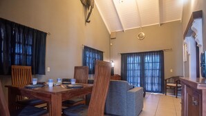 A dining area with a wooden table and chairs, a grey sofa, and a kitchen area with a clock on the wall. - NEW Casa Kudawecha (Noord)