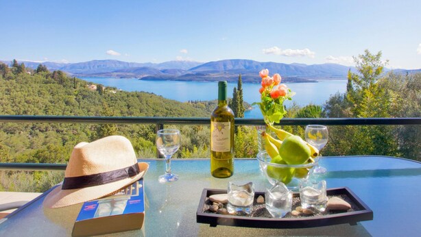 A table with a bottle of wine, glasses, a hat, and a tray with fruits and nuts, set against a scenic view of a lake and mountains.
