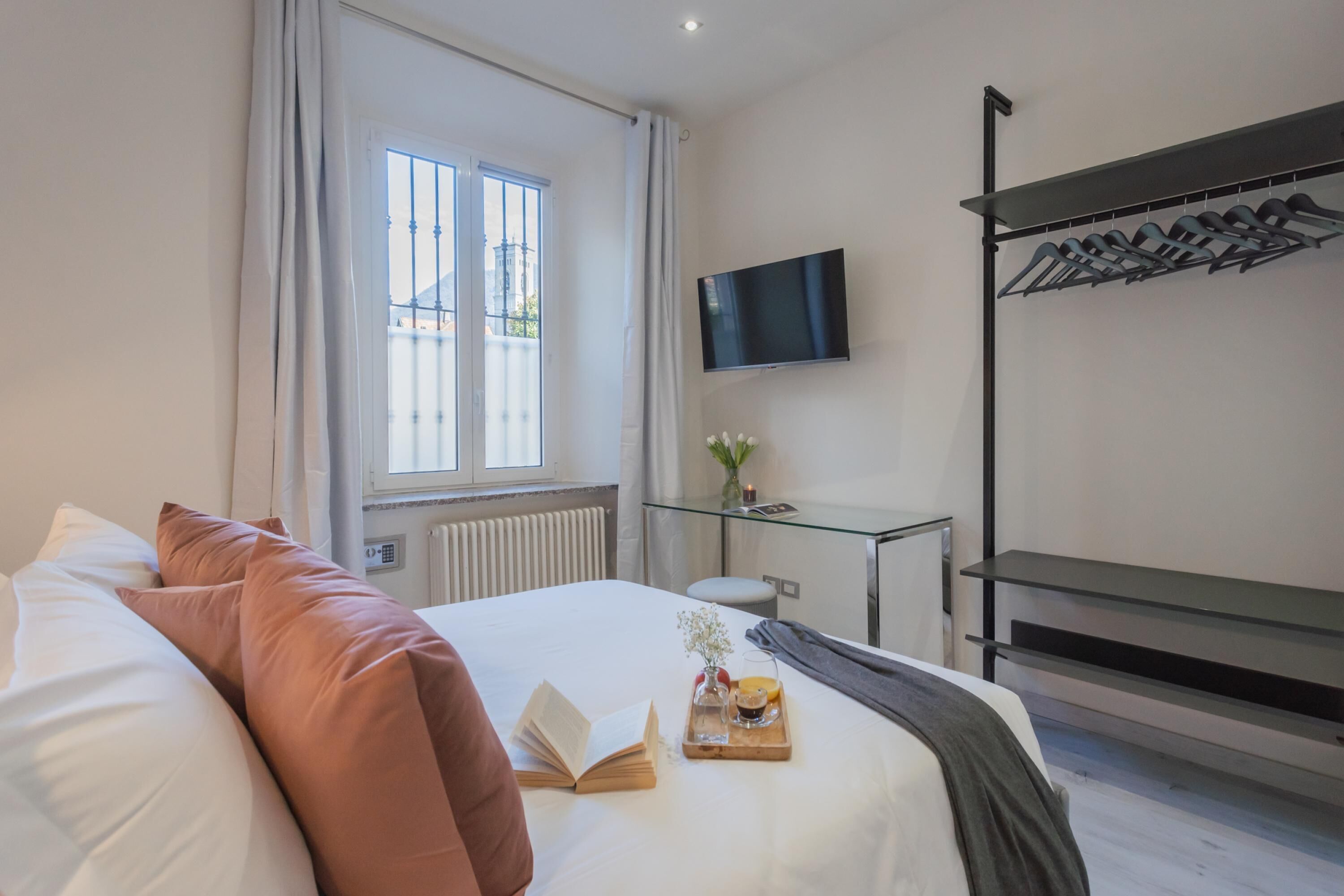 A hotel room with a bed, a TV mounted on the wall, a glass table with a tray of breakfast, and a window with curtains.