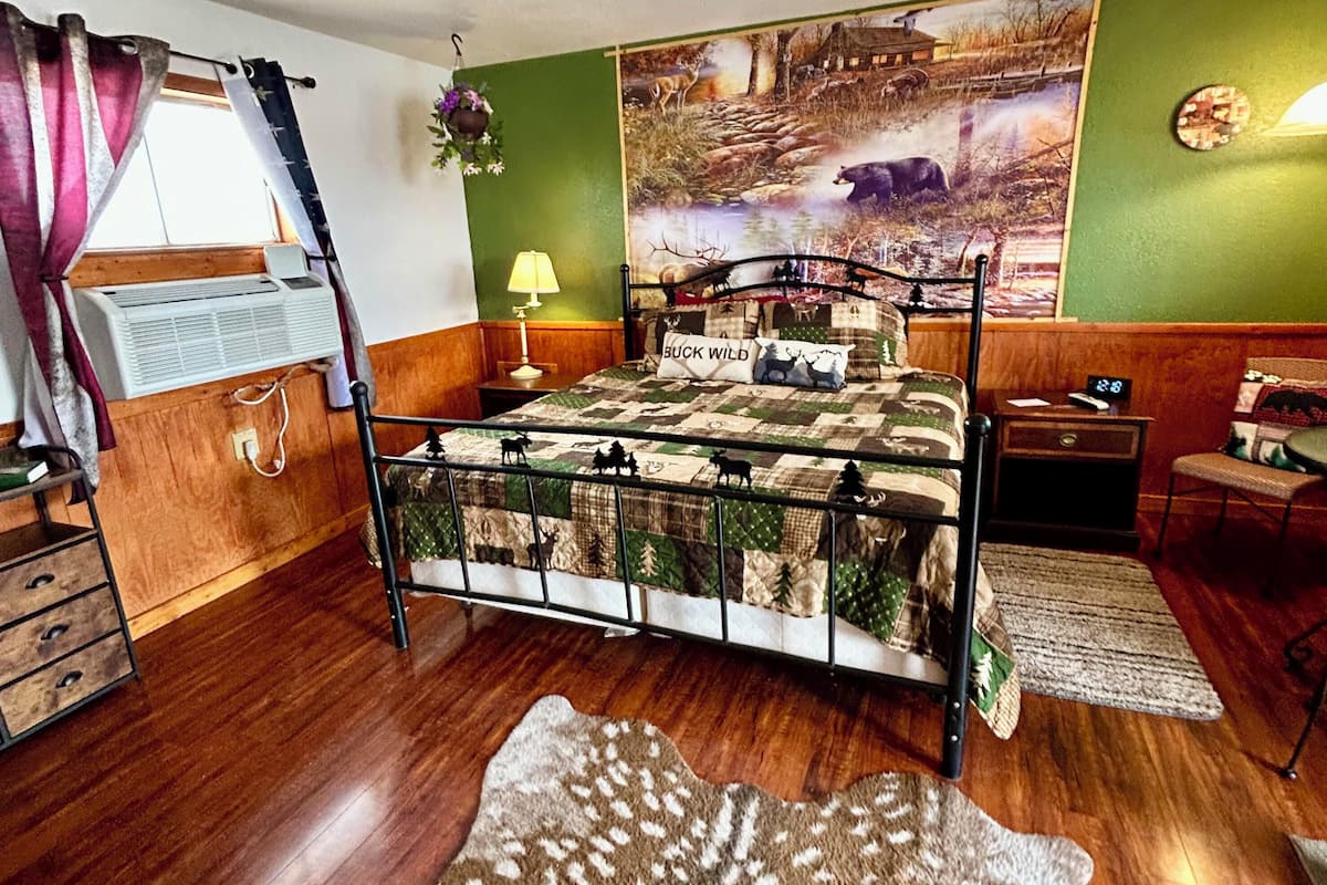 A rustic-themed bedroom with a bed, a wooden dresser, a nightstand, a lamp, and a large wall painting.